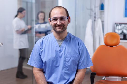 Close up portrait of orthodontist in dental office with patients in the background. Stomatolog in professioanl teeth clinic smiling wearing uniform looking at camera.