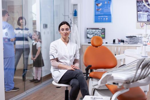 Ortodontist in dentistiry office sitting beside tools tray with assistant discussing with patietns in the background. Stomatolog in professioanl teeth clinic smiling wearing uniform looking at camera.