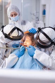 Mother looking at daughter with pain facial expression while dentist is treating caries. Stomatology team wearing ppe suit during covid19 doing procedure on child teeth.