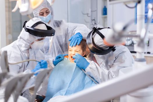 Mother comforting little girl with painfull expression in the course of dental treatment.Stomatology team wearing ppe suit during covid19 doing procedure on child teeth.