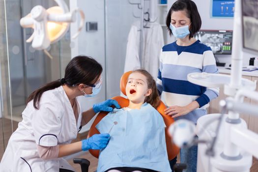 Dentist doing teeth check up of little girl sitting on dental chair wearing bib. Dentistry specialist during child cavity consultation in stomatology office using modern technology.