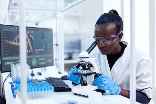 African doctor in pharmacology looking for sign of patoloy in patient sample using microscope. Black healthcare scientist in biochemistry laboratory wearing sterile equipment.