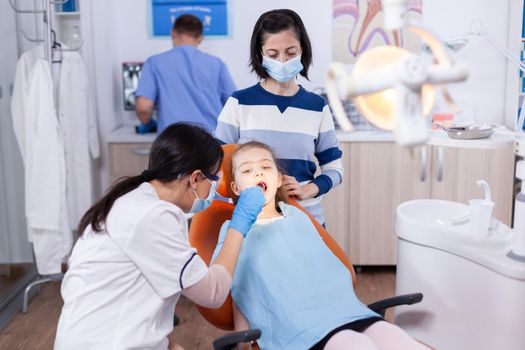 Stomatologist using curved mirror in the course of cavitiy examination of little girl wearing bib. Dentistry specialist during child cavity consultation in stomatology office using modern technology.