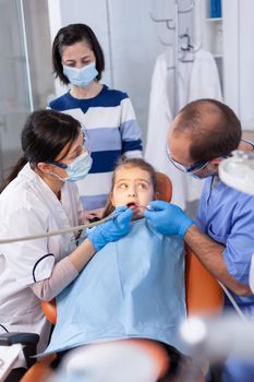 Little girl getting treatment for caries from dentist and assistant wearing dental bib. Mother with her kid in stomatology clinic for teeth examine using modern instruments.