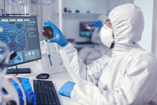 Medical scientist holding petri dish studying virus sample dressed in bacteriological suit Doctors analysing vaccine evolution using high tech researching diagnosis against covid19 virus.