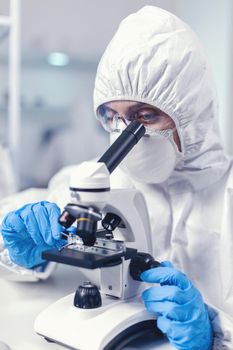 Woman scientis in coverall doing coronavirus investigation looking through microscope. Chemist researcher during global pandemic with covid-19 checking sample in biochemistry lab