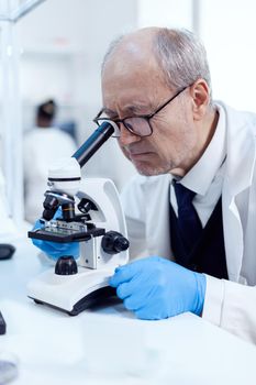 Senior scientist adjusting microscope while looking at virus and african assistant sitting at workplace. Chemist researcher in sterile lab doing experiments for medical industry using modern technology.