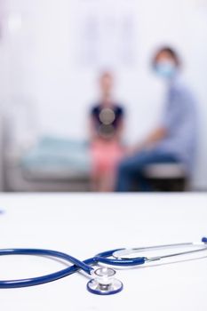 Close up of stethoscope in hospital office during coronavirus and nurse consulting child. Health doctor specialist providing health care services consultations treatment in protective equipment.