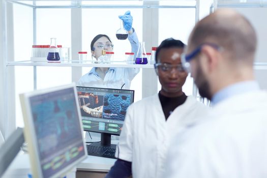 Expert chimist examining liquid in glass recipient. Multiethnic team of medical researchers working together in sterile lab wearing protection glasses and gloves.