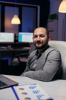 Focused freelancer smiling at camera resting hand on desk late at night. Smart businessman sitting at his workplace in the course of late night hours doing his job.