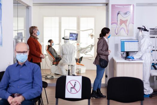 Patients with protection mask waiting in stomatology reception with new normal, staff wearing ppe suit. Woman checking appoiment with nurse coming for dental control during coronavirus pandemic