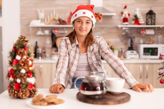 Portrait of child wearing santa hat while looking into camera enjoying winter season celebrating christmas holiday in xmas decorated culinary kitchen.. Baked x-mas cookies with coffee on table