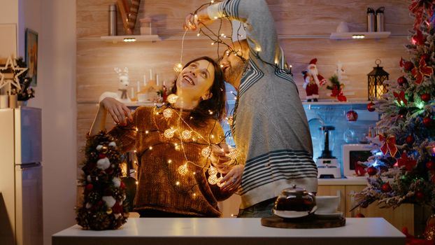 Cheerful people getting tangled in festive lights while decorating home for christmas eve celebration. Man and woman smiling, trying to untangle garland of twinkle lights bulbs.