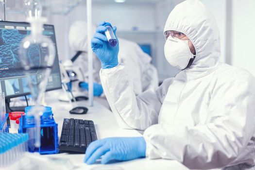 Lab technician dressed in protective suit as safety precaution looking at test tube. Scientist in laboratory wearing coverall doing research and analyzing substance during global pandemic with covid19.