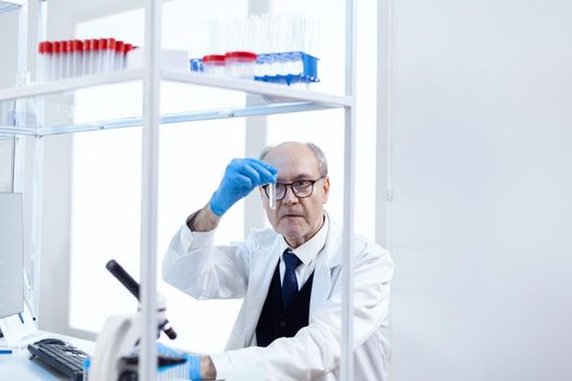 Male senior scientist in sterile medical laboratory looking at test tube wearing protection gear. Viorolog researcher in professional lab working to discover medical treatment.