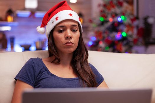 Female adult reading business email on laptop computer working during christmastime sitting comfortable on sofa in xmas decorated kitchen. Woman enjoying winter holiday celebrating christmas season