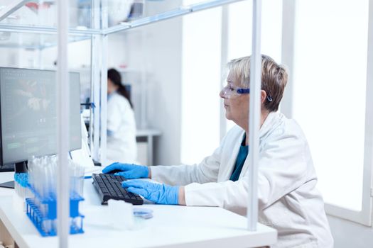 Elderly aged researcher working on computer sitting on her workplace. Senior scientist in pharmaceuticals laboratory doing genetic research wearing lab coat with team in the background.