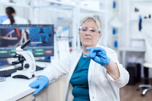 Senior scientist in protective gear holding sample during clinic study giving expertise. Elderly researcher in sterile lab looking on microscope slide wearing lab coat.