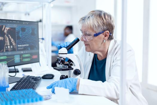 Senior scientist analysing sample of genetic material using microscope and african coworker in the background. Elderly researcher carrying out scientific research in a sterile lab with a modern technology.