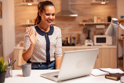 Cheerful woman waving during a video call while working late at night from home kitchen. Employee using modern technology at midnight doing overtime for job, business, career, network, lifestyle.