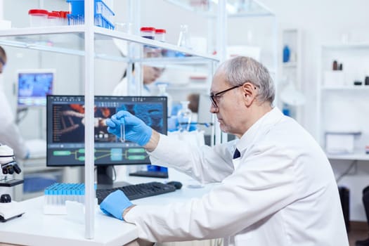 Senior scientist wearing protective gloves holding test tube in busy laboratory. Viorolog researcher in professional lab working to discover medical treatment.