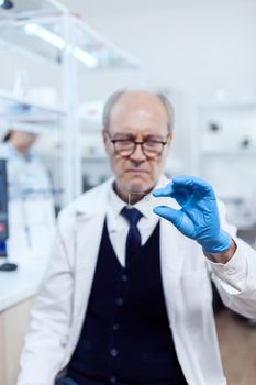 Experienced scientist in busy laboratory holding glass slide in microbiology laboratory. Senior researcher in sterile lab looking on microscope slide wearing lab coat.