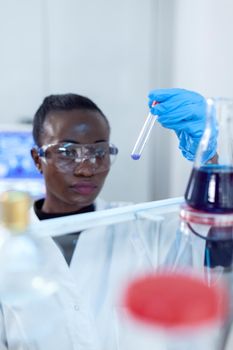 African scientist working with blue liquid in chemical test tubes wearing eyewear protection. Black researcher in sterile laboratory conducting pharmacology experiment wearing coat.
