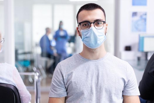 Young man with face mask against coronavirus looking at camera in hospital waiting area. Senior woman with walking frame waiting for consultation in clinic.