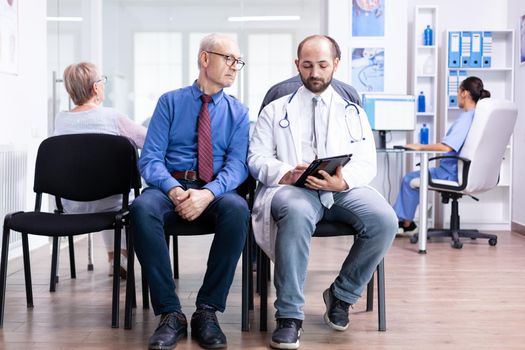 Doctor explaining test results to senior man in hospital waiting area. Disabled senior woman with walking frame arriving for medical examination.