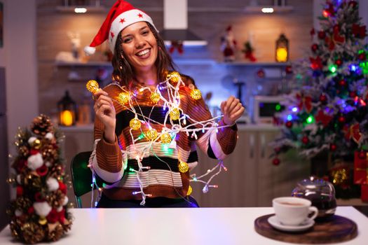 Caucasian woman decorating christmas tree with lights on string for holiday in festive kitchen at home. Young smiling person knotted in bright tangled garland wire illuminated bulbs