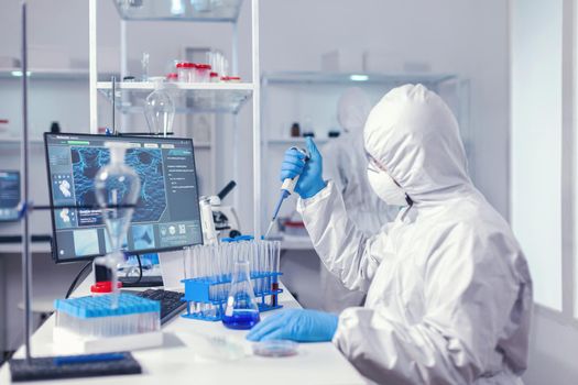 Laborant working with automatic pipette and test tubes dressed in protection suit. Chemist in modern laboratory doing research using dispenser during global epidemic with covid-19.