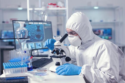 Biochemist testing sample for coronavirus using microscope dressed in ppe suit. Virolog in coverall during coronavirus outbreak conducting healthcare scientific analysis.