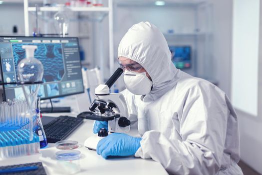 Healthcare specialist analyzing vaccine on microscope dressed in protection suit. Virolog in coverall during coronavirus outbreak conducting healthcare scientific analysis.