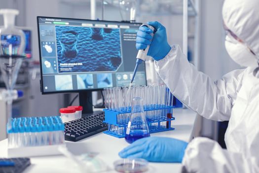 Laborant dropping sample in test tube using micropipette dressed in coverall. Chemist in modern laboratory doing research using dispenser during global epidemic with covid-19.