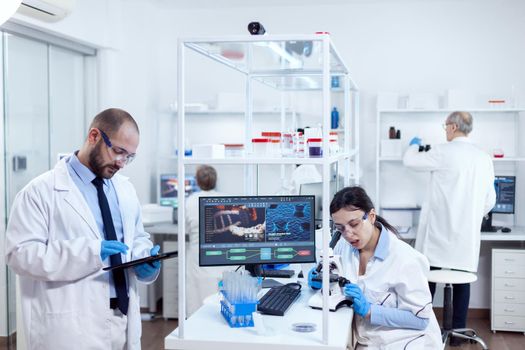 Group of scientist doing health study using microscope and tablet pc. Team of researchers doing pharmacology engineering in sterile laboratory for healthcare industry with african assistant in the background.