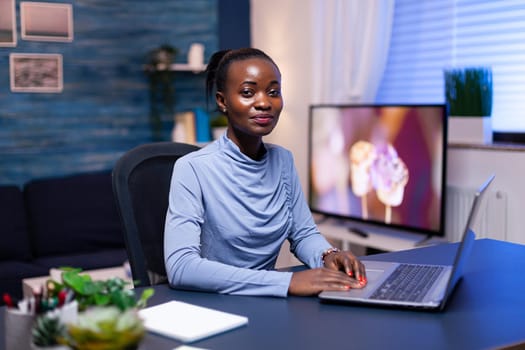 Successful african woman using laptop computer in living room late at night looking at camrea. Black entrepreneur sitting in personal workplace writing on keyboard.