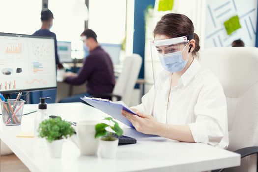 Financial advisor with wireless headphones checking financial statistic in busy office with face mask. Employees with visors working in corporate company workspace respecting social distance analysing data and graphs.