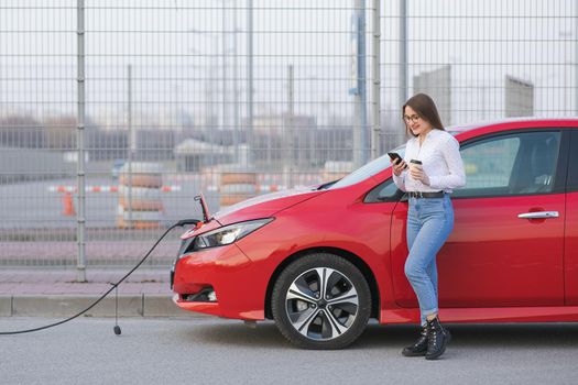 Girl Use Coffee Drink While Using Smart Phone and Waiting Power Supply Connect to Electric Vehicles for Charging the Battery in Car