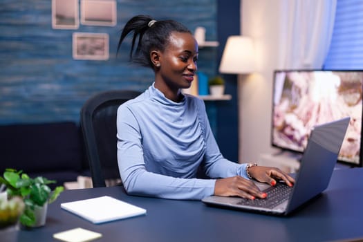 Dark skinned business woman working hard to finish a deadline using laptop computer from home late at night. Black entrepreneur sitting in personal workplace writing on keyboard.