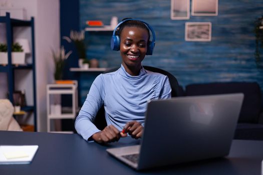 Black woman in good mood wearing headset listening music working on deadline from home office. sitting at desk. African freelancer creating new project working late.
