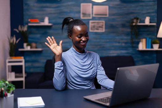 African business woman waving at clients in the course of virtual meeting sitting at desk in home office late at night. Black freelancer working with remotely team chatting virtual online conference.