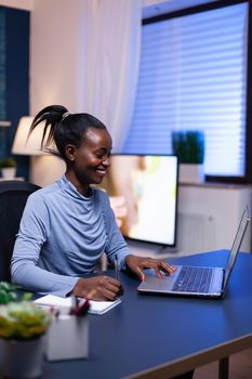 Dark skinned freelancer in the course of webinar smiling while taking notes sitting at desk late at night. Black entrepreneur sitting in personal workplace writing on keyboard.