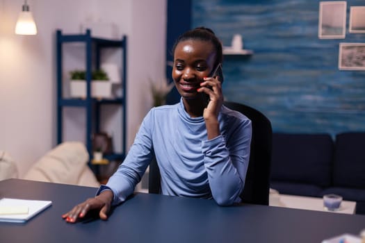 African business woman speaking on smartphone working late at night from home office. Busy focused freelancer using modern technology network wireless doing overtime.