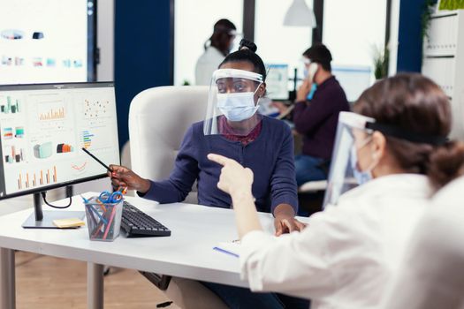 African manager explaining project graph to employee wearing face mask. Multiethnic team working in company with new normal respecting social distance because of global pandemic with coronavirus.