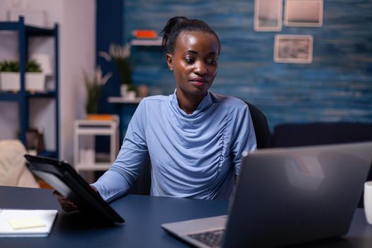 Concentrated african woman working on deadline using tablet pc and laptop in home office late at night. Busy focused employee using modern technology network wireless doing overtime writing.