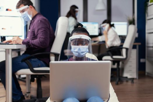 African freelancer in workplace using laptop wearing face mask against covid19. Multiethnic business team working respecting social distance during global pandemic with coronavirus.