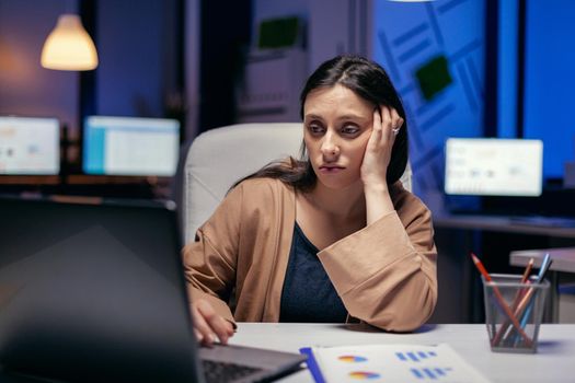 Lady in corporate office looking tired working on laptop doing overtime. Smart woman sitting at her workplace in the course of late night hours doing her job.