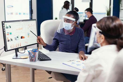 African businesswoman analysing colorful graph at offic wearing face mask. Multiethnic team working in company with new normal respecting social distance because of global pandemic with coronavirus.