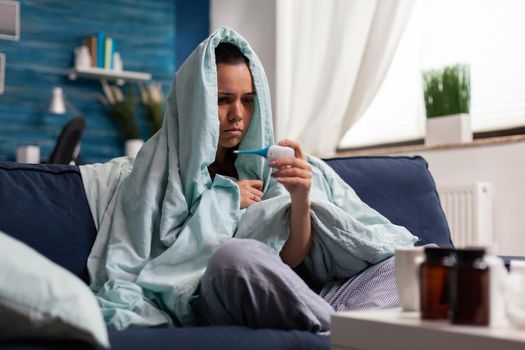 Caucasian woman measuring temperature ill at home with a thermometer. Person feeling sick cold unwell, checking fever and symptoms of disease flu infection. Resting adult with headache