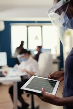 African bussinesswoman looking at graph and using tablet pc wearing face mask against covid19. Multiethnic coworkers respecting social distance in business company during global pandemic with coronavirus.